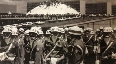 Fotografia em preto e branco. Em primeiro plano, um grupo de militares ocupa a parte inferior da imagem. Eles vestem uniformes escuros, capacetes brancos, cintos e bandoleiras brancas. Usam também máscaras ou lenços cobrindo o rosto. Ao fundo, há um grande número de pessoas reunidas em frente e sobre a cúpula branca do Congresso Nacional, em Brasília. Atrás da cúpula, aparecem os prédios retangulares das duas torres do Congresso. Diversos automóveis antigos estão estacionados ao longo da via em frente ao prédio. O céu está parcialmente nublado.