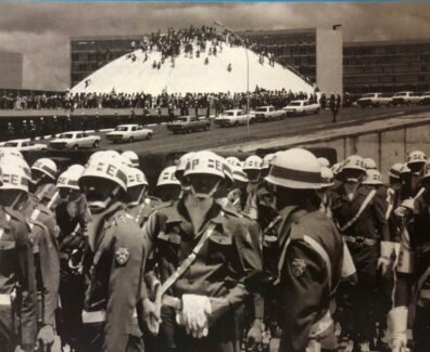 Fotografia em preto e branco. Em primeiro plano, um grupo de militares ocupa a parte inferior da imagem. Eles vestem uniformes escuros, capacetes brancos, cintos e bandoleiras brancas. Usam também máscaras ou lenços cobrindo o rosto. Ao fundo, há um grande número de pessoas reunidas em frente e sobre a cúpula branca do Congresso Nacional, em Brasília. Atrás da cúpula, aparecem os prédios retangulares das duas torres do Congresso. Diversos automóveis antigos estão estacionados ao longo da via em frente ao prédio. O céu está parcialmente nublado.