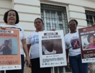 A imagem é um registro de um ato público em que três mulheres aparecem segurando cartazes. A manifestação, provavelmente realizada em Salvador (Bahia), é um protesto que clama por Justiça para jovens e crianças vítimas de violência armada, especialmente em casos que envolvem a polícia. As mulheres, que aparentam ser mães ou familiares das vítimas, estão paradas em frente a um prédio antigo, que tem uma fachada clássica e janelas gradeadas. A iluminação sugere que o protesto ocorreu ao entardecer ou em um dia nublado. ​Os cartazes, que são o foco da imagem, contêm o símbolo do Instituto Odara e do Fundo Baobá para Equidade Racial, confirmando que a manifestação é organizada por grupos de direitos humanos e familiares em busca de responsabilização do Estado pelos crimes. Foto: Jamile Novaes / Instituto Odara.