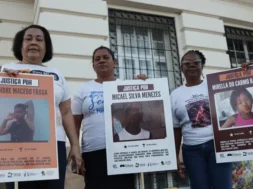 A imagem é um registro de um ato público em que três mulheres aparecem segurando cartazes. A manifestação, provavelmente realizada em Salvador (Bahia), é um protesto que clama por Justiça para jovens e crianças vítimas de violência armada, especialmente em casos que envolvem a polícia. As mulheres, que aparentam ser mães ou familiares das vítimas, estão paradas em frente a um prédio antigo, que tem uma fachada clássica e janelas gradeadas. A iluminação sugere que o protesto ocorreu ao entardecer ou em um dia nublado. Os cartazes, que são o foco da imagem, contêm o símbolo do Instituto Odara e do Fundo Baobá para Equidade Racial, confirmando que a manifestação é organizada por grupos de direitos humanos e familiares em busca de responsabilização do Estado pelos crimes. Foto: Jamile Novaes / Instituto Odara.