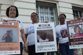 A imagem é um registro de um ato público em que três mulheres aparecem segurando cartazes. A manifestação, provavelmente realizada em Salvador (Bahia), é um protesto que clama por Justiça para jovens e crianças vítimas de violência armada, especialmente em casos que envolvem a polícia. As mulheres, que aparentam ser mães ou familiares das vítimas, estão paradas em frente a um prédio antigo, que tem uma fachada clássica e janelas gradeadas. A iluminação sugere que o protesto ocorreu ao entardecer ou em um dia nublado. ​Os cartazes, que são o foco da imagem, contêm o símbolo do Instituto Odara e do Fundo Baobá para Equidade Racial, confirmando que a manifestação é organizada por grupos de direitos humanos e familiares em busca de responsabilização do Estado pelos crimes. Foto: Jamile Novaes / Instituto Odara.