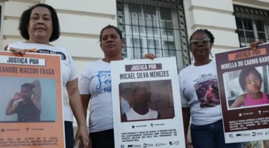 A imagem é um registro de um ato público em que três mulheres aparecem segurando cartazes. A manifestação, provavelmente realizada em Salvador (Bahia), é um protesto que clama por Justiça para jovens e crianças vítimas de violência armada, especialmente em casos que envolvem a polícia. As mulheres, que aparentam ser mães ou familiares das vítimas, estão paradas em frente a um prédio antigo, que tem uma fachada clássica e janelas gradeadas. A iluminação sugere que o protesto ocorreu ao entardecer ou em um dia nublado. ​Os cartazes, que são o foco da imagem, contêm o símbolo do Instituto Odara e do Fundo Baobá para Equidade Racial, confirmando que a manifestação é organizada por grupos de direitos humanos e familiares em busca de responsabilização do Estado pelos crimes. Foto: Jamile Novaes / Instituto Odara.