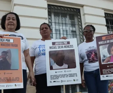 A imagem é um registro de um ato público em que três mulheres aparecem segurando cartazes. A manifestação, provavelmente realizada em Salvador (Bahia), é um protesto que clama por Justiça para jovens e crianças vítimas de violência armada, especialmente em casos que envolvem a polícia. As mulheres, que aparentam ser mães ou familiares das vítimas, estão paradas em frente a um prédio antigo, que tem uma fachada clássica e janelas gradeadas. A iluminação sugere que o protesto ocorreu ao entardecer ou em um dia nublado. Os cartazes, que são o foco da imagem, contêm o símbolo do Instituto Odara e do Fundo Baobá para Equidade Racial, confirmando que a manifestação é organizada por grupos de direitos humanos e familiares em busca de responsabilização do Estado pelos crimes. Foto: Jamile Novaes / Instituto Odara.