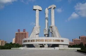 Na imagem tem três mãos em estátua, uma segurando um martelo simbolizando os trabalhadores da cidade, na esquerda, atrás segurando um pincel representando os intelectuais, e na direita a foice, representando os trabalhadores do campo.