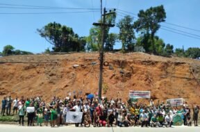 Moradores reunidos à frente de um morro completamente desmatado, segurando faixas, cartazes e bandeiras. Foto: UJC Brasil / Núcleo DC-SJM.