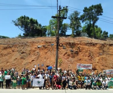 Moradores reunidos à frente de um morro completamente desmatado, segurando faixas, cartazes e bandeiras. Foto: UJC Brasil / Núcleo DC-SJM.