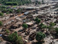 A imagem mostra uma paisagem devastada pela lama do rompimento da Barragem do Fundão, em Bento Rodrigues (Mariana–MG). Casas estão parcialmente soterradas ou destruídas, cobertas por uma espessa camada de rejeitos de mineração. Árvores aparecem retorcidas e encharcadas de lama, e um carro está preso entre os escombros. O cenário é de completa destruição: o mar de lama ocupa todo o espaço, substituindo o antigo distrito por um ambiente desolado, onde se misturam terra, entulho e silêncio.