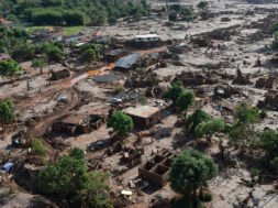 A imagem mostra uma paisagem devastada pela lama do rompimento da Barragem do Fundão, em Bento Rodrigues (Mariana–MG). Casas estão parcialmente soterradas ou destruídas, cobertas por uma espessa camada de rejeitos de mineração. Árvores aparecem retorcidas e encharcadas de lama, e um carro está preso entre os escombros. O cenário é de completa destruição: o mar de lama ocupa todo o espaço, substituindo o antigo distrito por um ambiente desolado, onde se misturam terra, entulho e silêncio.