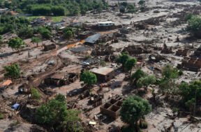 A imagem mostra uma paisagem devastada pela lama do rompimento da Barragem do Fundão, em Bento Rodrigues (Mariana–MG). Casas estão parcialmente soterradas ou destruídas, cobertas por uma espessa camada de rejeitos de mineração. Árvores aparecem retorcidas e encharcadas de lama, e um carro está preso entre os escombros. O cenário é de completa destruição: o mar de lama ocupa todo o espaço, substituindo o antigo distrito por um ambiente desolado, onde se misturam terra, entulho e silêncio.
