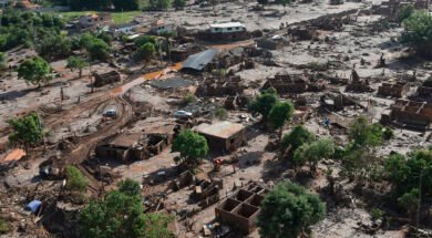A imagem mostra uma paisagem devastada pela lama do rompimento da Barragem do Fundão, em Bento Rodrigues (Mariana–MG). Casas estão parcialmente soterradas ou destruídas, cobertas por uma espessa camada de rejeitos de mineração. Árvores aparecem retorcidas e encharcadas de lama, e um carro está preso entre os escombros. O cenário é de completa destruição: o mar de lama ocupa todo o espaço, substituindo o antigo distrito por um ambiente desolado, onde se misturam terra, entulho e silêncio.