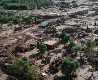 A imagem mostra uma paisagem devastada pela lama do rompimento da Barragem do Fundão, em Bento Rodrigues (Mariana–MG). Casas estão parcialmente soterradas ou destruídas, cobertas por uma espessa camada de rejeitos de mineração. Árvores aparecem retorcidas e encharcadas de lama, e um carro está preso entre os escombros. O cenário é de completa destruição: o mar de lama ocupa todo o espaço, substituindo o antigo distrito por um ambiente desolado, onde se misturam terra, entulho e silêncio.