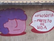 Foto de um muro de concreto com rachaduras verticais e manchas de desgaste. No centro da imagem, há um grafite composto por dois elementos: à esquerda, um desenho de rosto estilizado com formato redondo, pele rosa e cabelo roxo; à direita, um balão de fala branco com contorno vermelho. Dentro do balão, em letras vermelhas, está escrito: “+ FILOSOFIA – PROJETO DE VIDA”. Arte: @souls_ujc