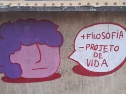 Foto de um muro de concreto com rachaduras verticais e manchas de desgaste. No centro da imagem, há um grafite composto por dois elementos: à esquerda, um desenho de rosto estilizado com formato redondo, pele rosa e cabelo roxo; à direita, um balão de fala branco com contorno vermelho. Dentro do balão, em letras vermelhas, está escrito: “+ FILOSOFIA – PROJETO DE VIDA”. Arte: @souls_ujc