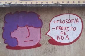 Foto de um muro de concreto com rachaduras verticais e manchas de desgaste. No centro da imagem, há um grafite composto por dois elementos: à esquerda, um desenho de rosto estilizado com formato redondo, pele rosa e cabelo roxo; à direita, um balão de fala branco com contorno vermelho. Dentro do balão, em letras vermelhas, está escrito: “+ FILOSOFIA – PROJETO DE VIDA”. Arte: @souls_ujc
