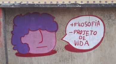 Foto de um muro de concreto com rachaduras verticais e manchas de desgaste. No centro da imagem, há um grafite composto por dois elementos: à esquerda, um desenho de rosto estilizado com formato redondo, pele rosa e cabelo roxo; à direita, um balão de fala branco com contorno vermelho. Dentro do balão, em letras vermelhas, está escrito: “+ FILOSOFIA – PROJETO DE VIDA”. Arte: @souls_ujc