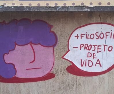 Foto de um muro de concreto com rachaduras verticais e manchas de desgaste. No centro da imagem, há um grafite composto por dois elementos: à esquerda, um desenho de rosto estilizado com formato redondo, pele rosa e cabelo roxo; à direita, um balão de fala branco com contorno vermelho. Dentro do balão, em letras vermelhas, está escrito: “+ FILOSOFIA – PROJETO DE VIDA”. Arte: @souls_ujc