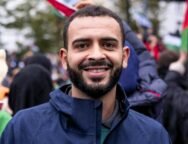 Foto de Mohammed Khatib, um homem palestino de 36 anos. Mohammed tem pele marrom, usa uma barba preenchida e veste um casaco. Ao fundo, uma manifestação com bandeiras da Palestina.