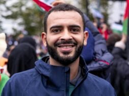 Foto de Mohammed Khatib, um homem palestino de 36 anos. Mohammed tem pele marrom, usa uma barba preenchida e veste um casaco. Ao fundo, uma manifestação com bandeiras da Palestina.