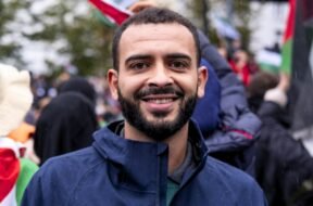 Foto de Mohammed Khatib, um homem palestino de 36 anos. Mohammed tem pele marrom, usa uma barba preenchida e veste um casaco. Ao fundo, uma manifestação com bandeiras da Palestina.