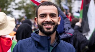 Foto de Mohammed Khatib, um homem palestino de 36 anos. Mohammed tem pele marrom, usa uma barba preenchida e veste um casaco. Ao fundo, uma manifestação com bandeiras da Palestina.