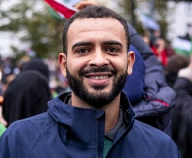 Foto de Mohammed Khatib, um homem palestino de 36 anos. Mohammed tem pele marrom, usa uma barba preenchida e veste um casaco. Ao fundo, uma manifestação com bandeiras da Palestina.