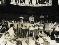 Fotografia em preto e branco de uma grande assembleia de estudantes sentados e em pé durante o Congresso da UBES de 1981; alguns falam ao microfone enquanto outros aplaudem, com uma faixa ao fundo defendendo a UBES e a organização do movimento estudantil.