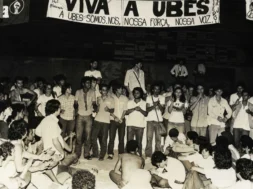 Fotografia em preto e branco de uma grande assembleia de estudantes sentados e em pé durante o Congresso da UBES de 1981; alguns falam ao microfone enquanto outros aplaudem, com uma faixa ao fundo defendendo a UBES e a organização do movimento estudantil.