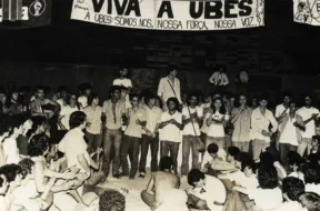 Fotografia em preto e branco de uma grande assembleia de estudantes sentados e em pé durante o Congresso da UBES de 1981; alguns falam ao microfone enquanto outros aplaudem, com uma faixa ao fundo defendendo a UBES e a organização do movimento estudantil.