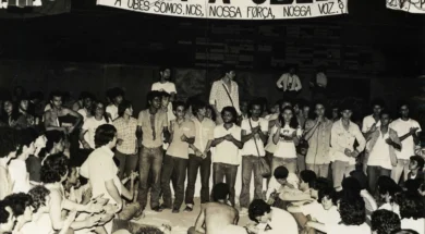 Fotografia em preto e branco de uma grande assembleia de estudantes sentados e em pé durante o Congresso da UBES de 1981; alguns falam ao microfone enquanto outros aplaudem, com uma faixa ao fundo defendendo a UBES e a organização do movimento estudantil.