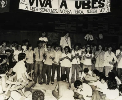 Fotografia em preto e branco de uma grande assembleia de estudantes sentados e em pé durante o Congresso da UBES de 1981; alguns falam ao microfone enquanto outros aplaudem, com uma faixa ao fundo defendendo a UBES e a organização do movimento estudantil.