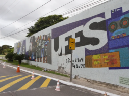 Fotografia de um painel de mosaico em um muro da universidade com a palavra “UFES” em letras grandes. O mural inclui ilustrações estilizadas de pessoas, elementos tecnológicos e gráficos coloridos. Em frente ao muro há uma rua com faixa de pedestres amarela e cones de sinalização, além de um poste e árvores ao fundo sob um céu nublado.