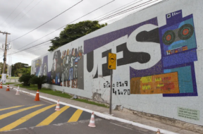 Fotografia de um painel de mosaico em um muro da universidade com a palavra “UFES” em letras grandes. O mural inclui ilustrações estilizadas de pessoas, elementos tecnológicos e gráficos coloridos. Em frente ao muro há uma rua com faixa de pedestres amarela e cones de sinalização, além de um poste e árvores ao fundo sob um céu nublado.