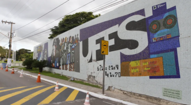 Fotografia de um painel de mosaico em um muro da universidade com a palavra “UFES” em letras grandes. O mural inclui ilustrações estilizadas de pessoas, elementos tecnológicos e gráficos coloridos. Em frente ao muro há uma rua com faixa de pedestres amarela e cones de sinalização, além de um poste e árvores ao fundo sob um céu nublado.