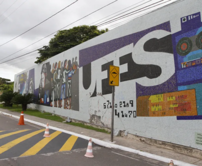 Fotografia de um painel de mosaico em um muro da universidade com a palavra “UFES” em letras grandes. O mural inclui ilustrações estilizadas de pessoas, elementos tecnológicos e gráficos coloridos. Em frente ao muro há uma rua com faixa de pedestres amarela e cones de sinalização, além de um poste e árvores ao fundo sob um céu nublado.
