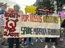 Registro fotográfico de militantes da população LGBTI+ com uma faixa branca escrito “LGBTI+ Tem classe, Por nossos direitos, saúde, moradia e trabalho” Outres ao fundo seguram cartazes que trazem mensagens de apoio à população LGBT e críticas à LGBTfobia. Foto: Lucas Nogueira / UJC Brasil.