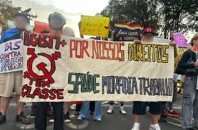 Registro fotográfico de militantes da população LGBTI+ com uma faixa branca escrito “LGBTI+ Tem classe, Por nossos direitos, saúde, moradia e trabalho” Outres ao fundo seguram cartazes que trazem mensagens de apoio à população LGBT e críticas à LGBTfobia. Foto: Lucas Nogueira / UJC Brasil.