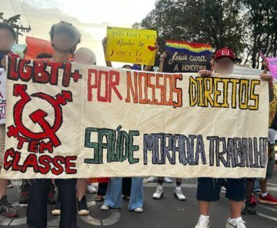 Registro fotográfico de militantes da população LGBTI+ com uma faixa branca escrito “LGBTI+ Tem classe, Por nossos direitos, saúde, moradia e trabalho” Outres ao fundo seguram cartazes que trazem mensagens de apoio à população LGBT e críticas à LGBTfobia. Foto: Lucas Nogueira / UJC Brasil.