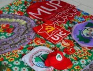 A imagem mostra uma mesa coberta por uma toalha florida colorida, sobre a qual estão dispostos materiais de movimentos populares e estudantis, como camisas, bonés, bandeiras e panfletos da União da Juventude Comunista (UJC), do Movimento por uma Universidade Popular (MUP), do Movimento de Mulheres Camponesas (MMC), e do Movimento dos Atingidos por Barragens (MAB), compondo um cenário de formação política, organização popular e articulação entre juventude, universidade e luta camponesa. Créditos: Movimento por uma Universidade Popular (MUP).