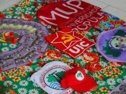 A imagem mostra uma mesa coberta por uma toalha florida colorida, sobre a qual estão dispostos materiais de movimentos populares e estudantis, como camisas, bonés, bandeiras e panfletos da União da Juventude Comunista (UJC), do Movimento por uma Universidade Popular (MUP), do Movimento de Mulheres Camponesas (MMC), e do Movimento dos Atingidos por Barragens (MAB), compondo um cenário de formação política, organização popular e articulação entre juventude, universidade e luta camponesa. Créditos: Movimento por uma Universidade Popular (MUP).