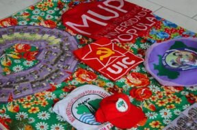 A imagem mostra uma mesa coberta por uma toalha florida colorida, sobre a qual estão dispostos materiais de movimentos populares e estudantis, como camisas, bonés, bandeiras e panfletos da União da Juventude Comunista (UJC), do Movimento por uma Universidade Popular (MUP), do Movimento de Mulheres Camponesas (MMC), e do Movimento dos Atingidos por Barragens (MAB), compondo um cenário de formação política, organização popular e articulação entre juventude, universidade e luta camponesa. Créditos: Movimento por uma Universidade Popular (MUP).