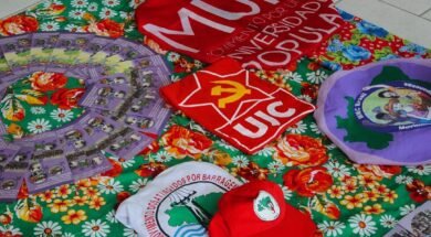 A imagem mostra uma mesa coberta por uma toalha florida colorida, sobre a qual estão dispostos materiais de movimentos populares e estudantis, como camisas, bonés, bandeiras e panfletos da União da Juventude Comunista (UJC), do Movimento por uma Universidade Popular (MUP), do Movimento de Mulheres Camponesas (MMC), e do Movimento dos Atingidos por Barragens (MAB), compondo um cenário de formação política, organização popular e articulação entre juventude, universidade e luta camponesa. Créditos: Movimento por uma Universidade Popular (MUP).