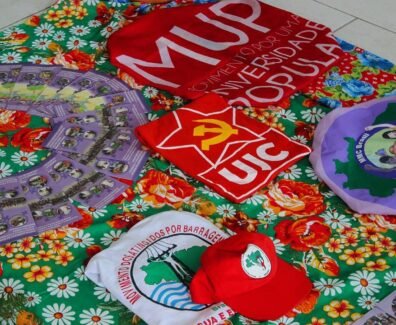 A imagem mostra uma mesa coberta por uma toalha florida colorida, sobre a qual estão dispostos materiais de movimentos populares e estudantis, como camisas, bonés, bandeiras e panfletos da União da Juventude Comunista (UJC), do Movimento por uma Universidade Popular (MUP), do Movimento de Mulheres Camponesas (MMC), e do Movimento dos Atingidos por Barragens (MAB), compondo um cenário de formação política, organização popular e articulação entre juventude, universidade e luta camponesa. Créditos: Movimento por uma Universidade Popular (MUP).