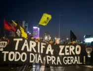 Fotografia de manifestação durante a noite. A imagem contém pessoas atrás de uma faixa preta com o texto "Tarifa Zero todo dia pra geral!" em branco, junto a desenhos de catracas em chamas nas laterais. Algumas pessoas movimentam bandeiras de organizações políticas. No fundo, há alguns edifícios.