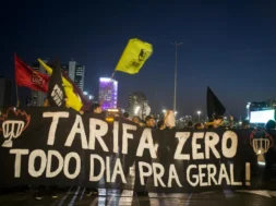 Fotografia de manifestação durante a noite. A imagem contém pessoas atrás de uma faixa preta com o texto "Tarifa Zero todo dia pra geral!" em branco, junto a desenhos de catracas em chamas nas laterais. Algumas pessoas movimentam bandeiras de organizações políticas. No fundo, há alguns edifícios.