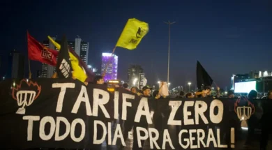 Fotografia de manifestação durante a noite. A imagem contém pessoas atrás de uma faixa preta com o texto "Tarifa Zero todo dia pra geral!" em branco, junto a desenhos de catracas em chamas nas laterais. Algumas pessoas movimentam bandeiras de organizações políticas. No fundo, há alguns edifícios.
