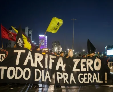 Fotografia de manifestação durante a noite. A imagem contém pessoas atrás de uma faixa preta com o texto "Tarifa Zero todo dia pra geral!" em branco, junto a desenhos de catracas em chamas nas laterais. Algumas pessoas movimentam bandeiras de organizações políticas. No fundo, há alguns edifícios.